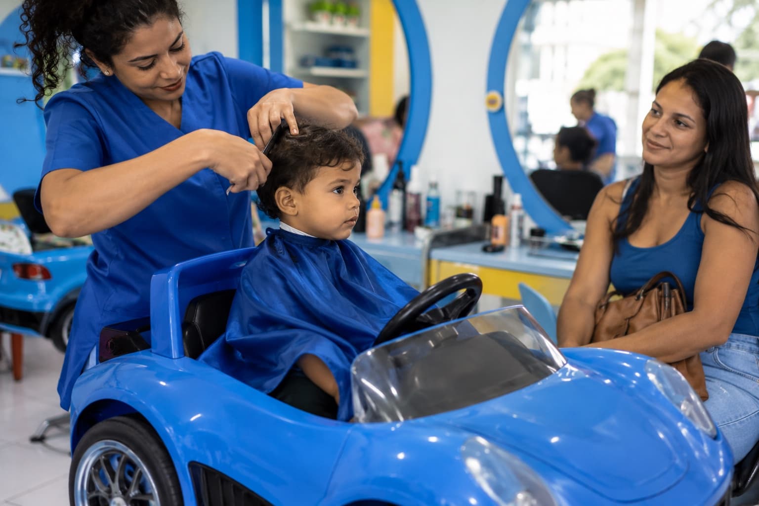 Criança sentada em cadeira em formato de carrinho cortando o cabelo com cabeleireira de azul, mãe observando