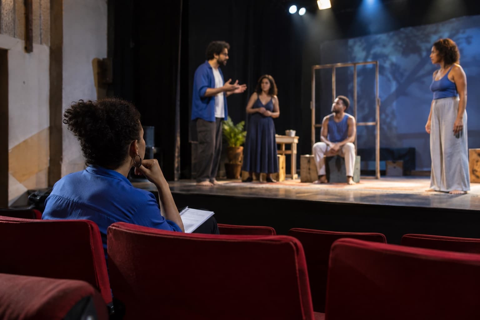 Interior de um pequeno teatro com atores ensaiando no palco sob holofotes enquanto o diretor de cena observa das poltronas vermelhas vazias
