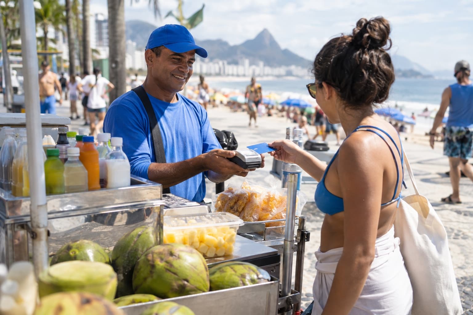 Vendedor ambulante de boné azul num calçadão de praia recebendo pagamento por cartão numa maquininha, ao lado de um carrinho com bebidas e petiscos