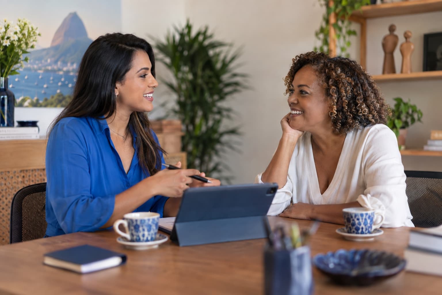 Consultora de blusa azul conversando com cliente de meia-idade em mesa de escritório aconchegante com café e tablet