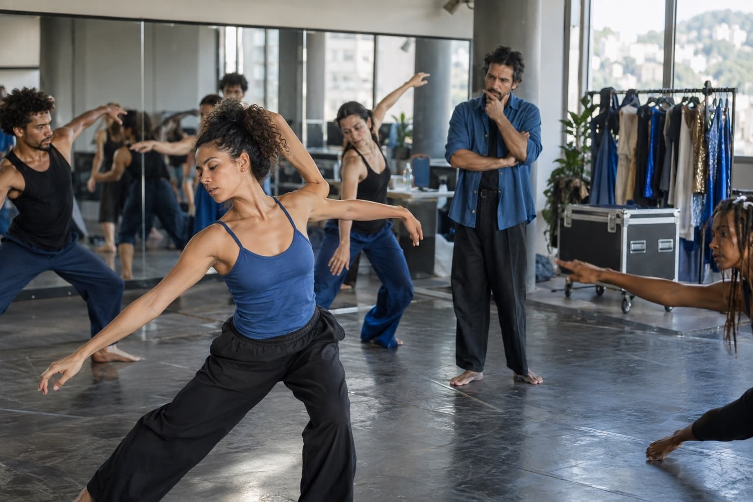 Companhia de dança contemporânea ensaiando em sala com espelhos, coreógrafa de azul observando e arara de figurinos ao lado