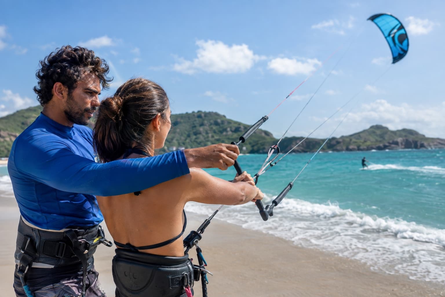 Instrutor de kitesurf de lycra azul ensinando aluno a segurar a barra de controle em praia ventosa com kite no ar e mar turquesa