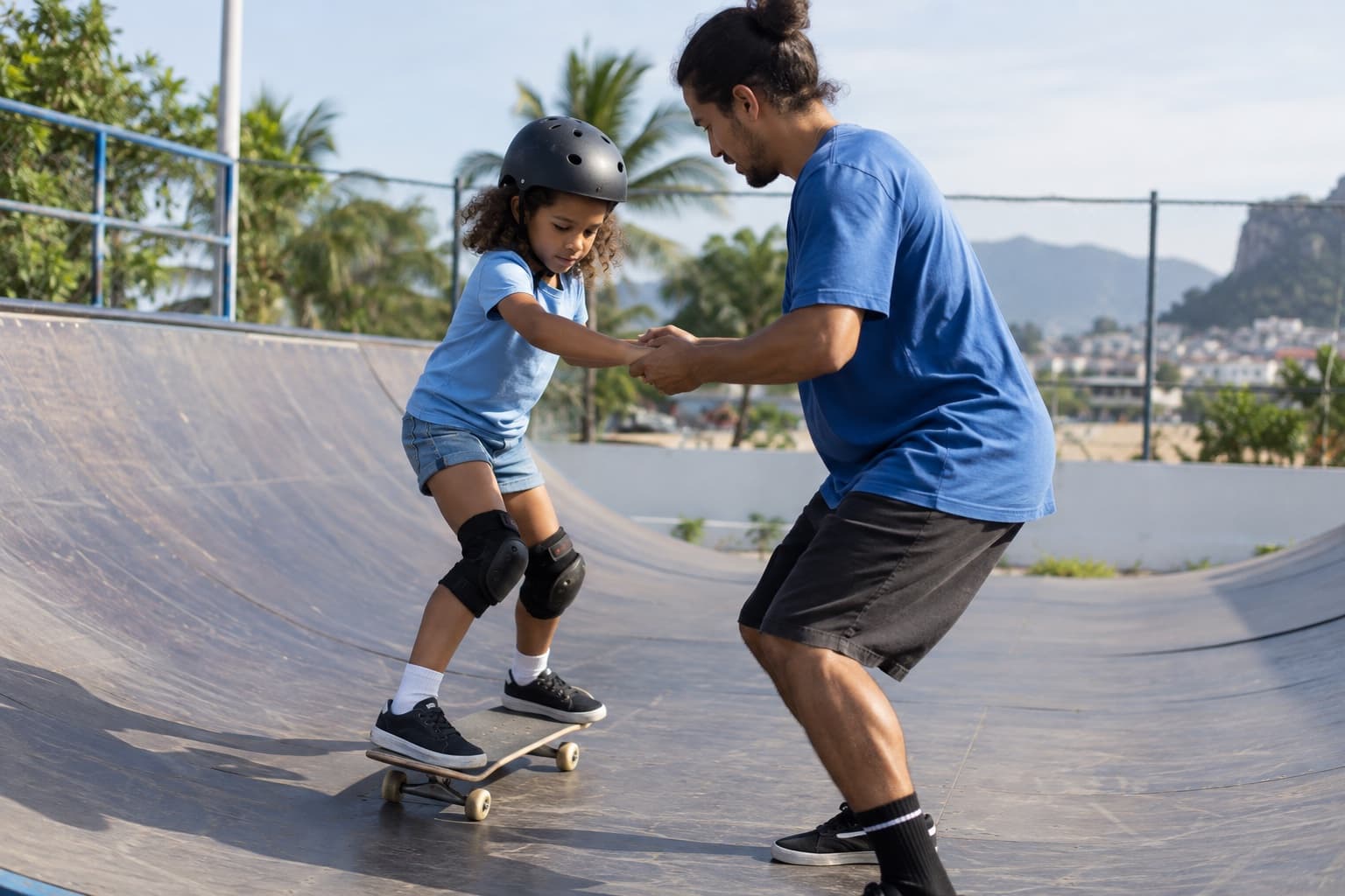 Instrutor de skate de camiseta azul segurando as mãos de uma criança com capacete e joelheiras andando de skate numa mini rampa de pista ao ar livre