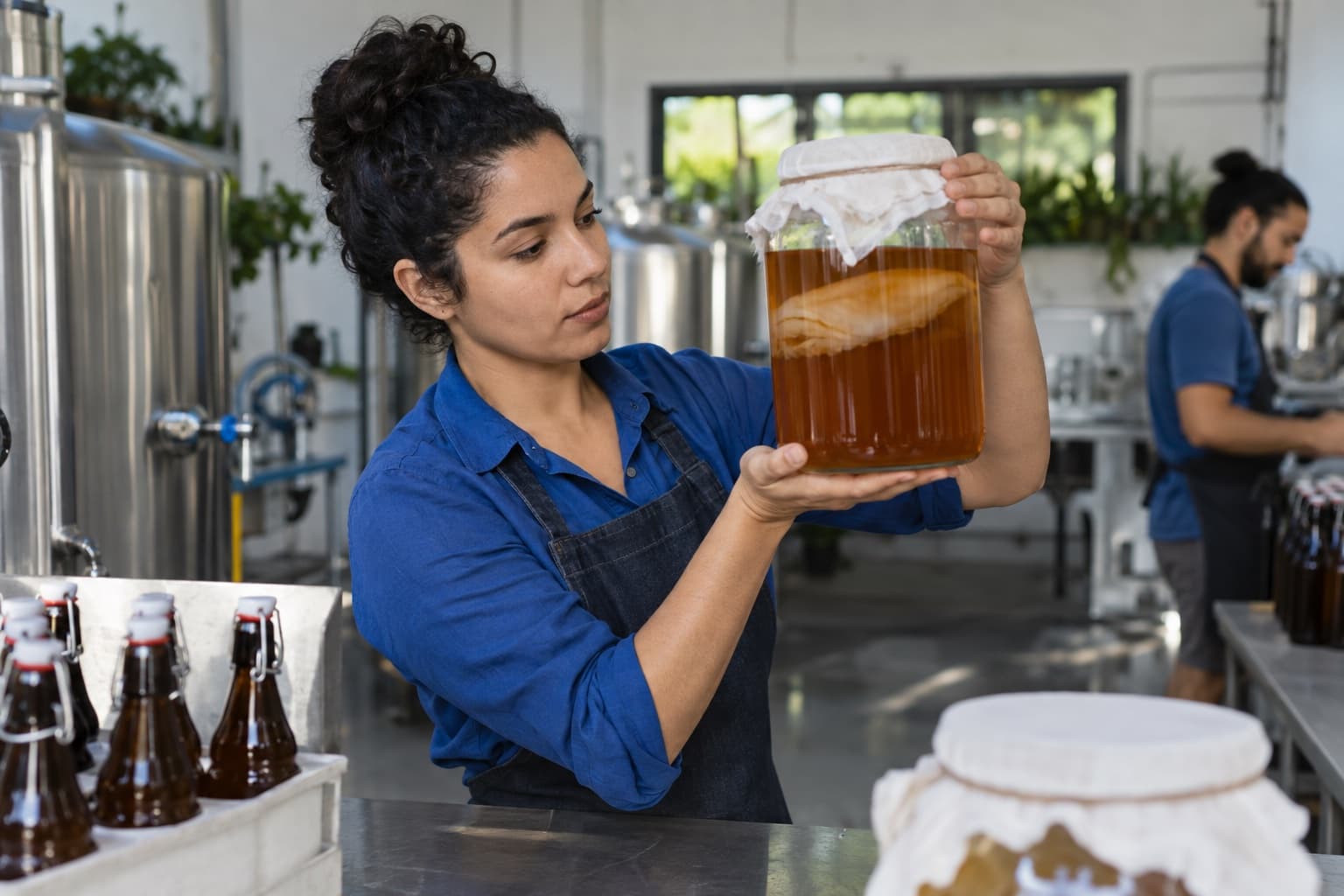Produtora de camisa azul conferindo um grande vidro de fermentação de kombucha com a cultura flutuando, ao lado de tanques de inox e garrafas numa pequena fábrica