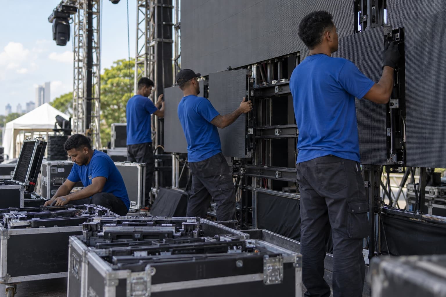 Técnicos de camiseta azul montando painéis de LED numa estrutura de treliça no palco de um evento, com cases abertos no chão