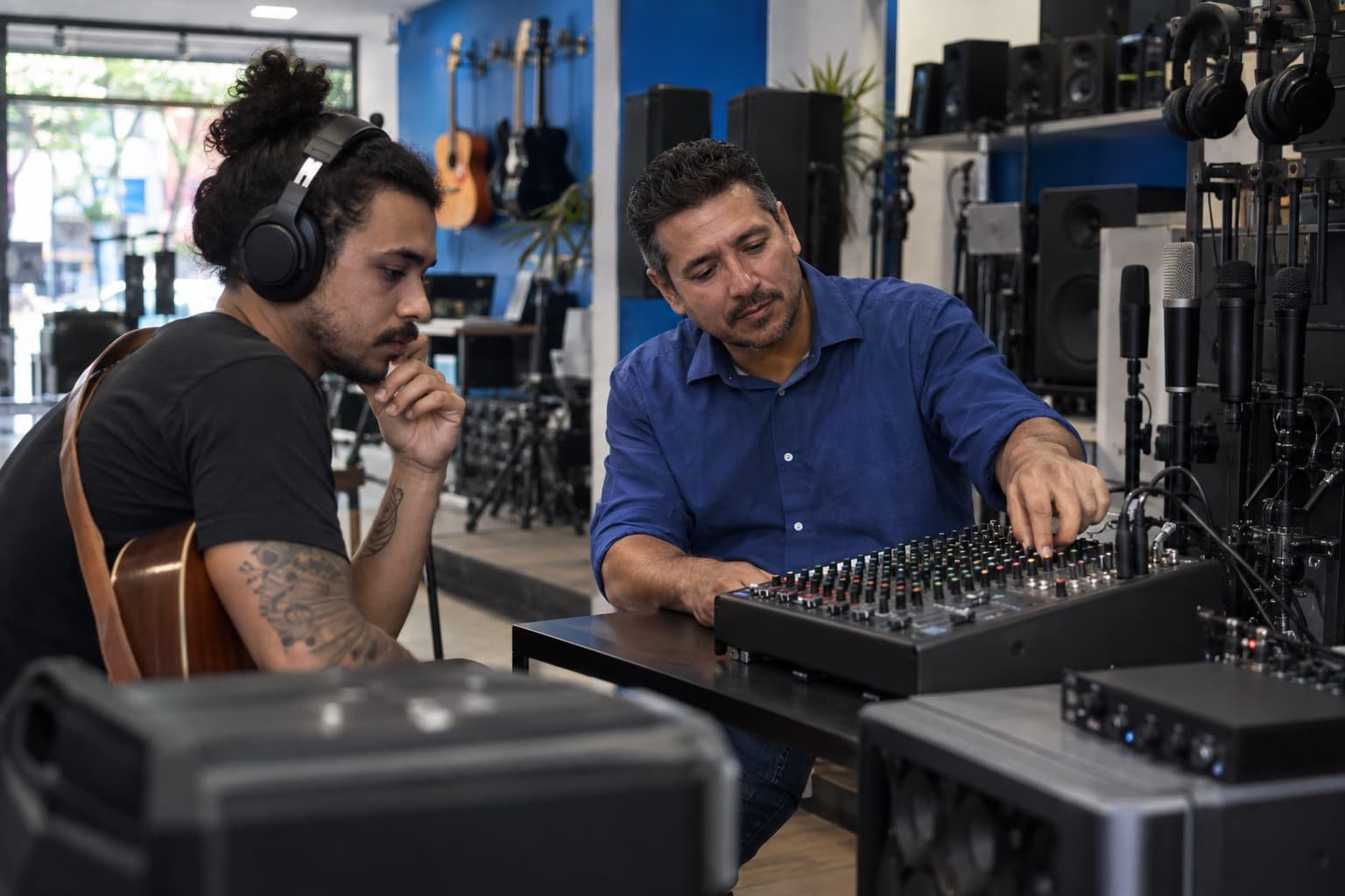 Loja de áudio profissional com mesas de som, caixas acústicas e microfones, vendedor de camisa azul testando uma mesa com fones de ouvido para um músico