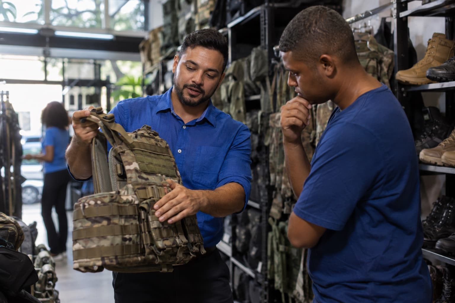 Loja de artigos táticos com mochilas, coturnos e coletes camuflados nas prateleiras, vendedor de camisa azul mostrando um colete a um cliente