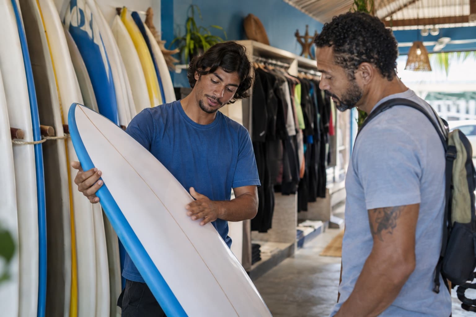 Interior de surf shop com pranchas em suportes e roupas de borracha penduradas, vendedor jovem de camiseta azul mostrando uma prancha a um cliente