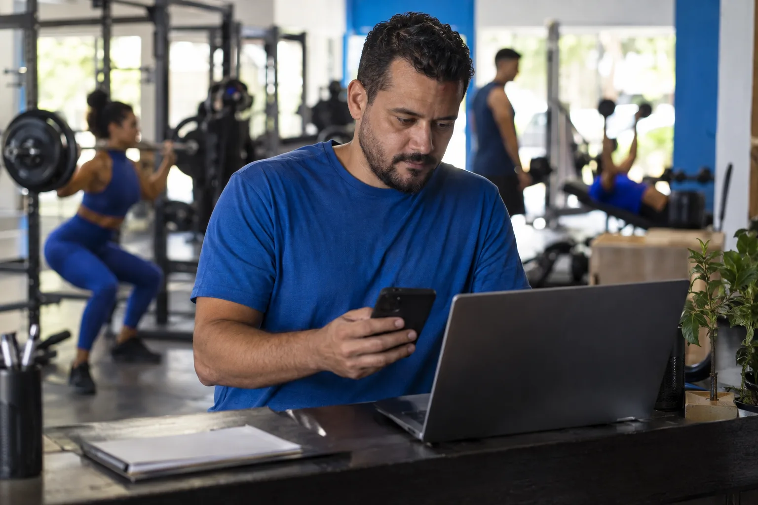 Dono de uma academia confere o celular e o notebook na recepção enquanto alunos treinam ao fundo