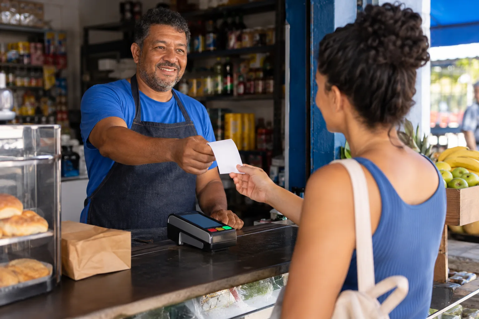 Dono de mercadinho entrega comprovante impresso à cliente no balcão ao lado da maquininha